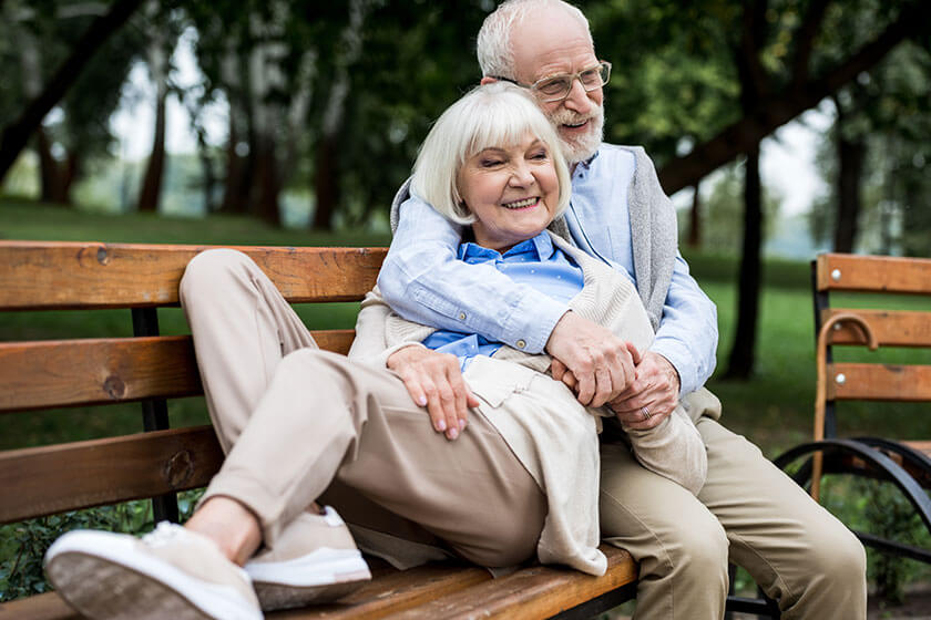 smiling senior woman laying on wooden bench near happy husband in park smiling senior woman laying on wooden bench near happy husband in park