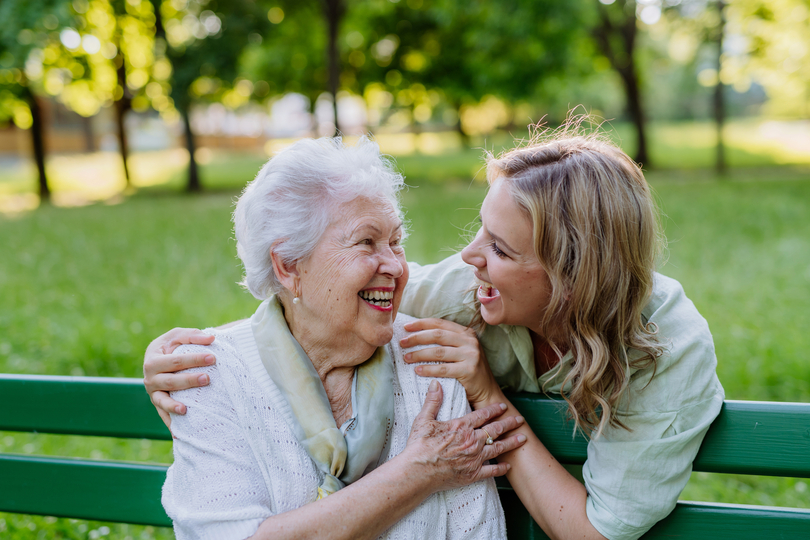 Adult granddaguhter helping her grandmother to use cellphone when sitting on bench in park in summer. Adult granddaguhter helping her grandmother to use cellphone when sitting on bench in park in summer.